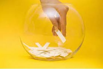 vaso in vetro trasparente su sfondo giallo, contenente bigliettini di colore bianco con una mano che estrae uno di quei biglietti